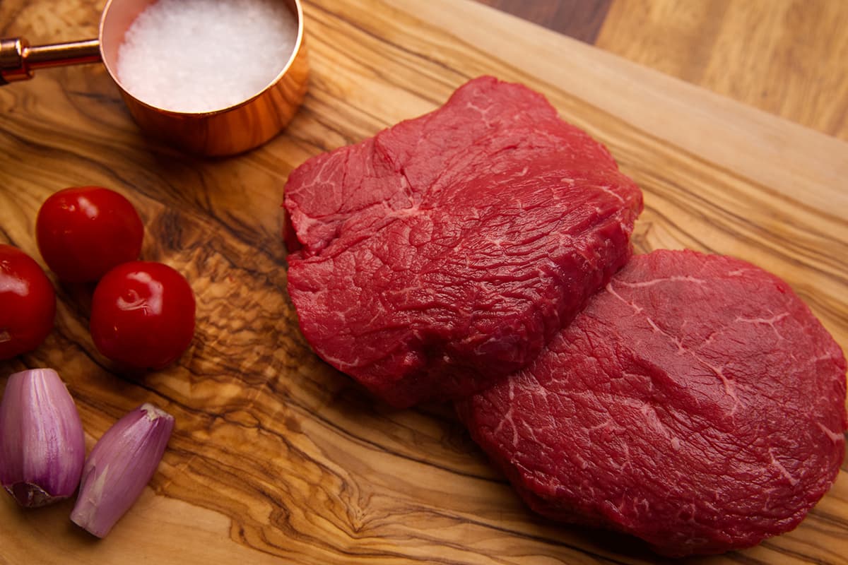 Close up of two Kingsbury Wagyu Fillet steaks on a chopping board with tomatoes, salt granules and shallots